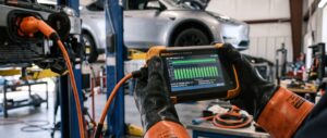 Kfz-Sachverständiger mit Hochvolt-Schutzhandschuhen wertet Messdaten auf einem Diagnosegerät aus – professionelle Batteriediagnose für Kfz-Gutachter an einem E-Auto auf der Hebebühne.